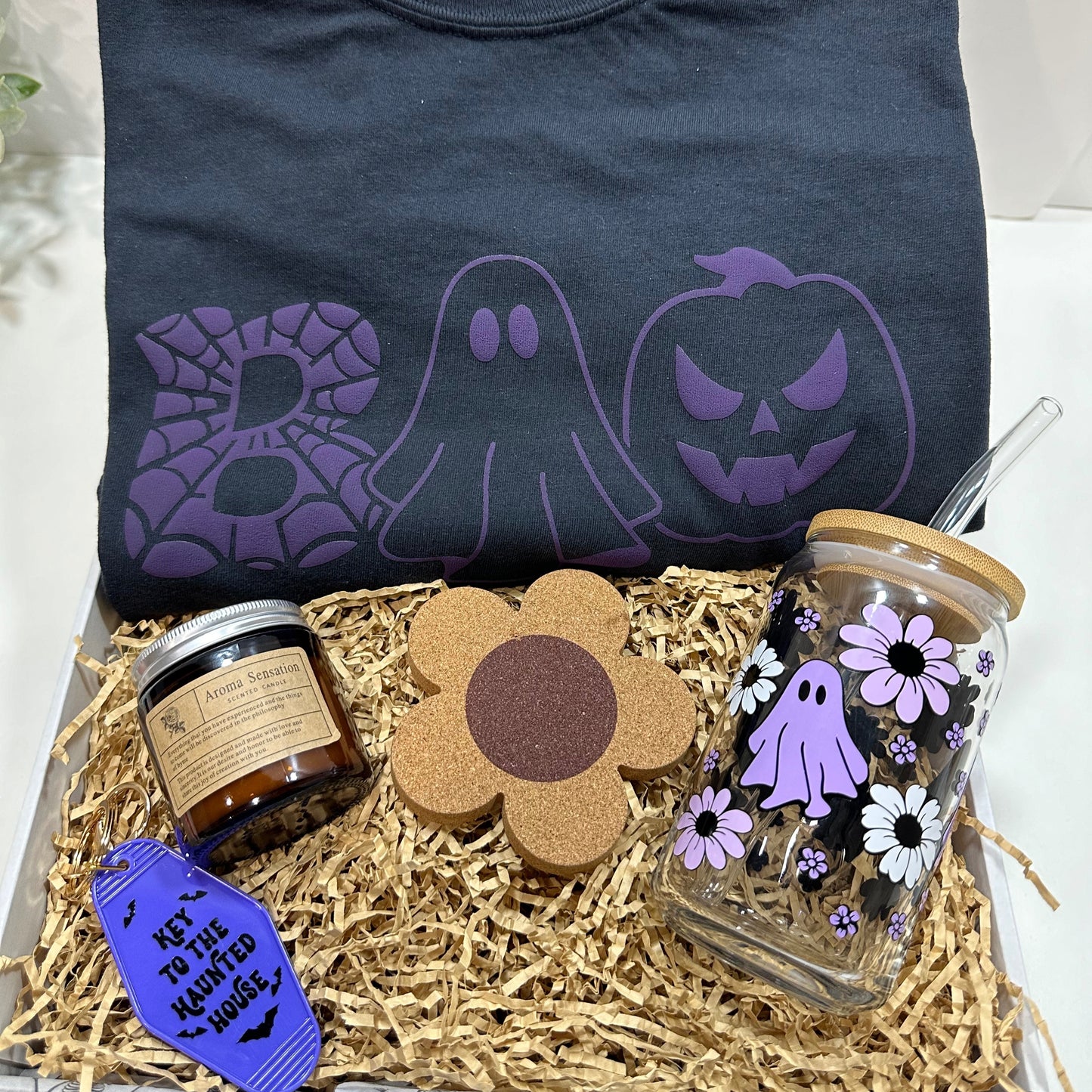 basket with black shirt that has a boo purple design , flower cork coaster, ghost and flowers glass can cup with lid and straw, a candle and a purple motel keychain that has bats and said key to the haunted house.