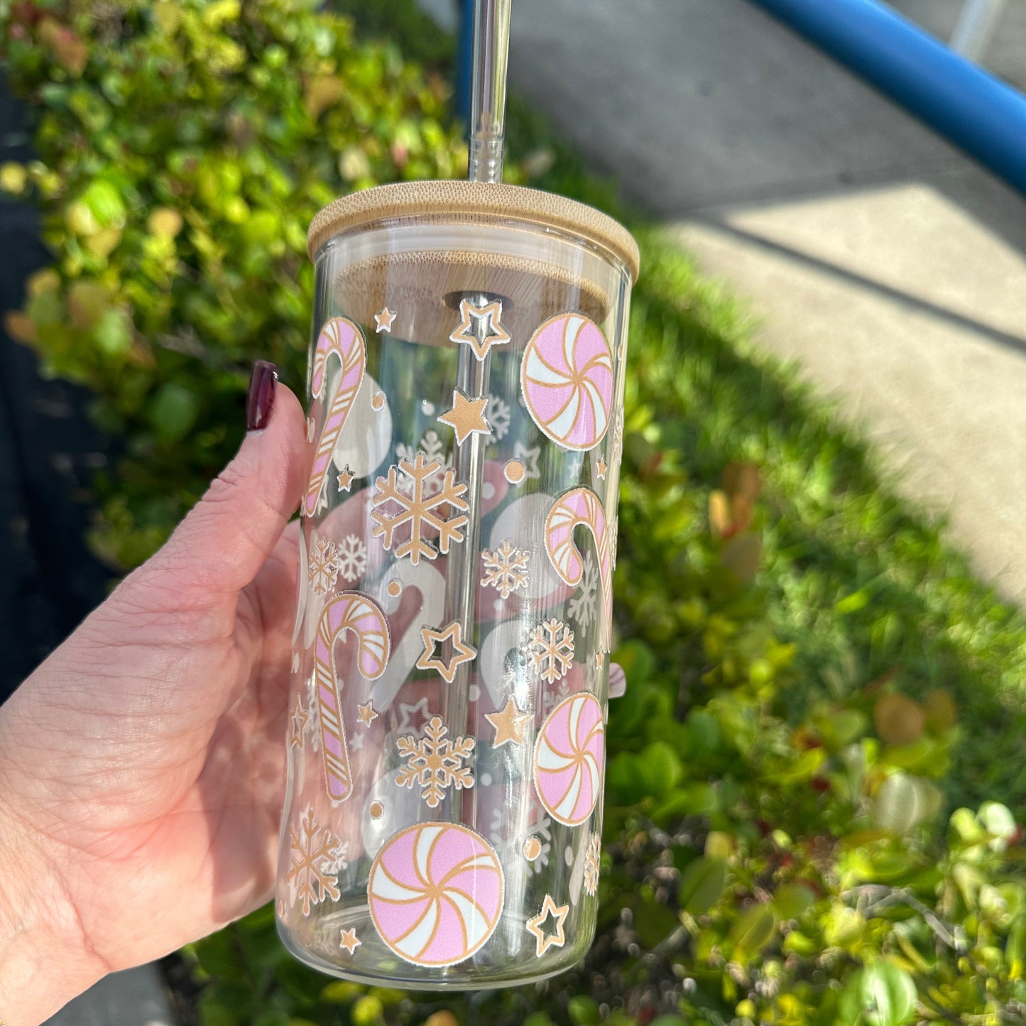 A glass cup with a candy cane pattern and stars, held by a person against a backdrop of greenery.