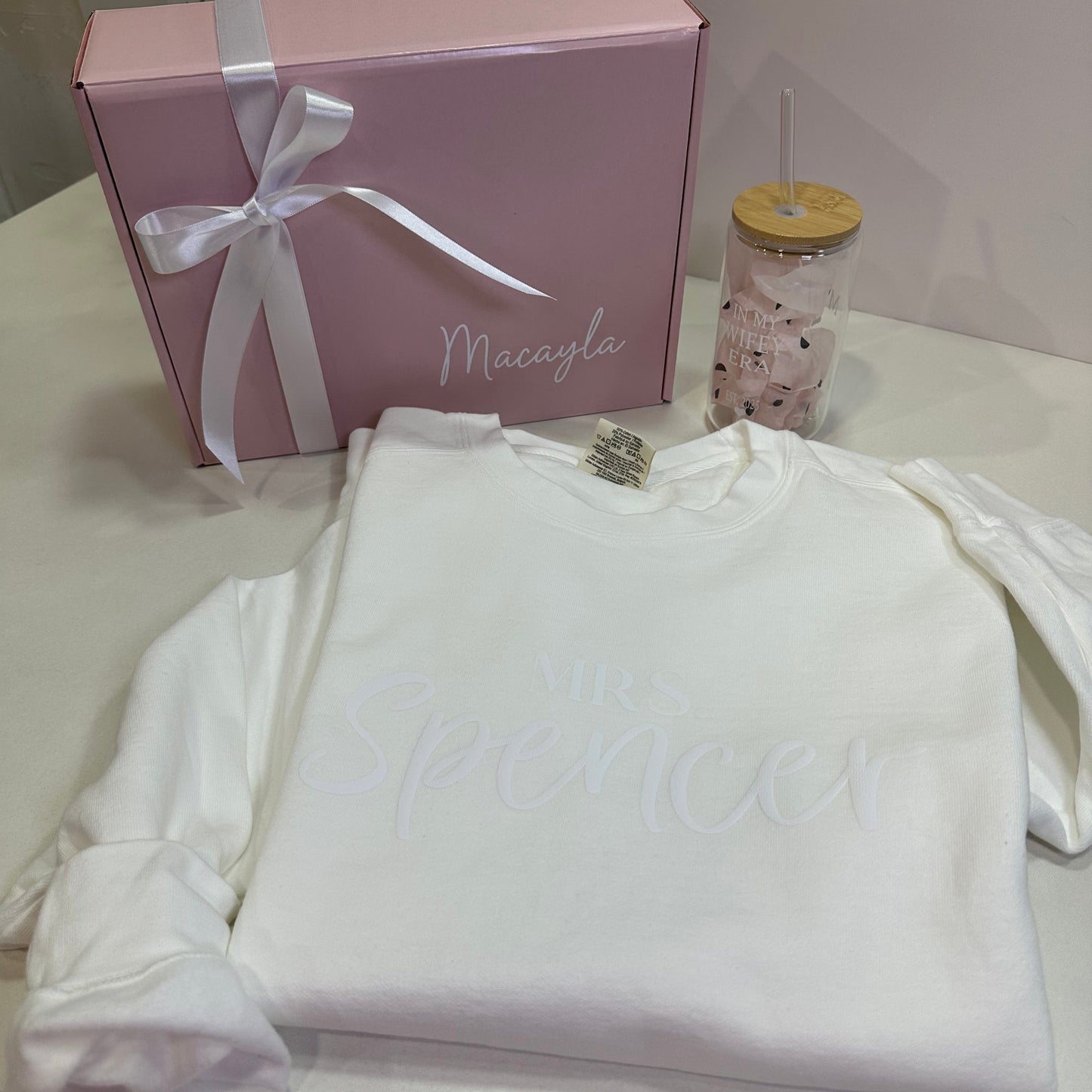 White mrs sweatshirt on a white surface next to a personalized pink gift box with a bow and a in my wifey era cup