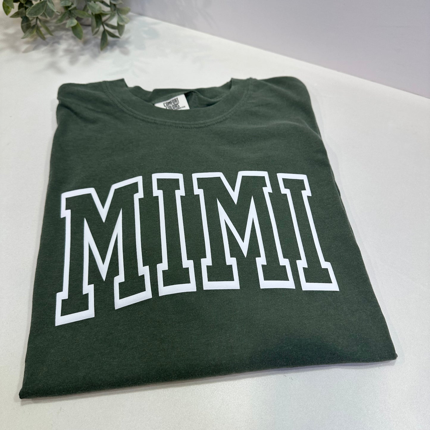 Green t-shirt with 'MIMI' printed on it, placed on a white surface.