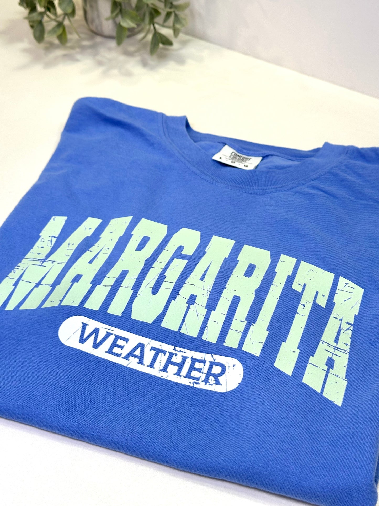 Blue t-shirt with 'Margarita Weather' text on a white surface