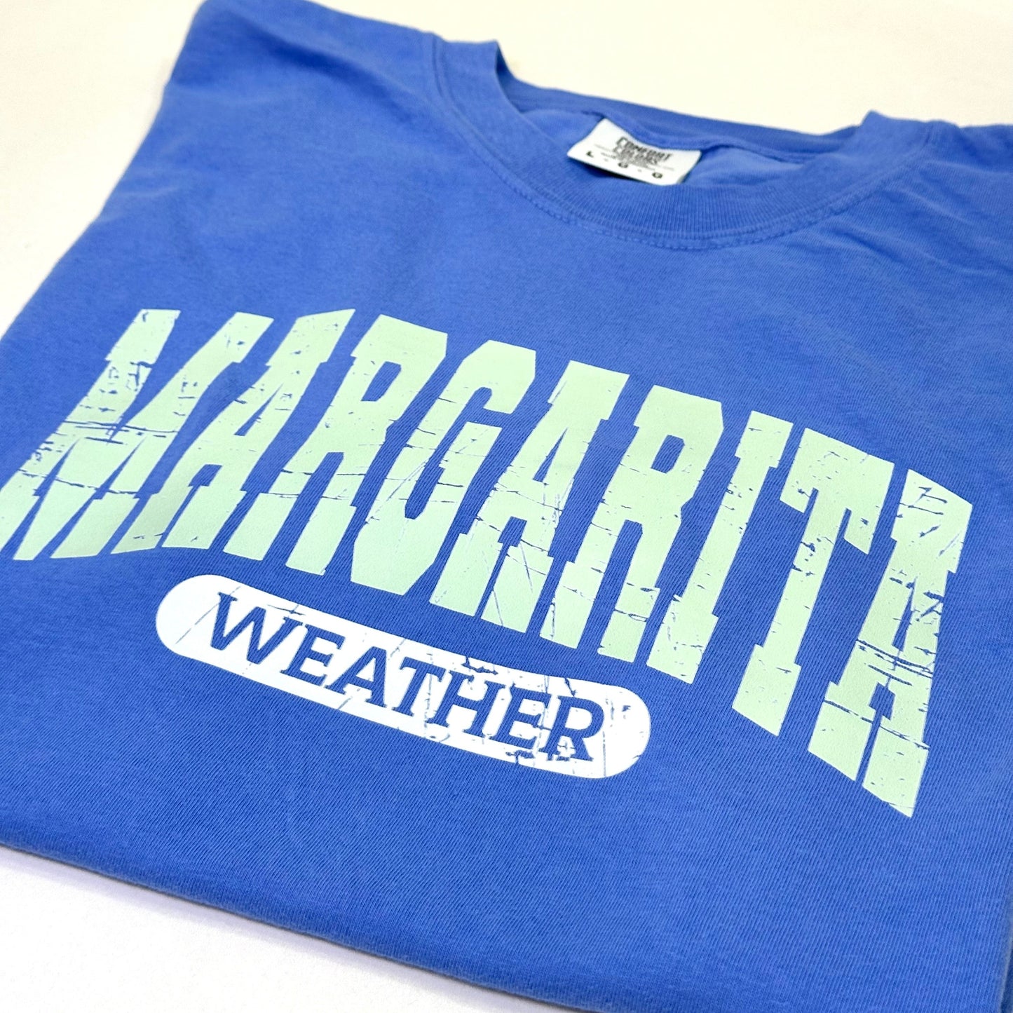 Blue t-shirt with 'Margarita Weather' text on a white surface