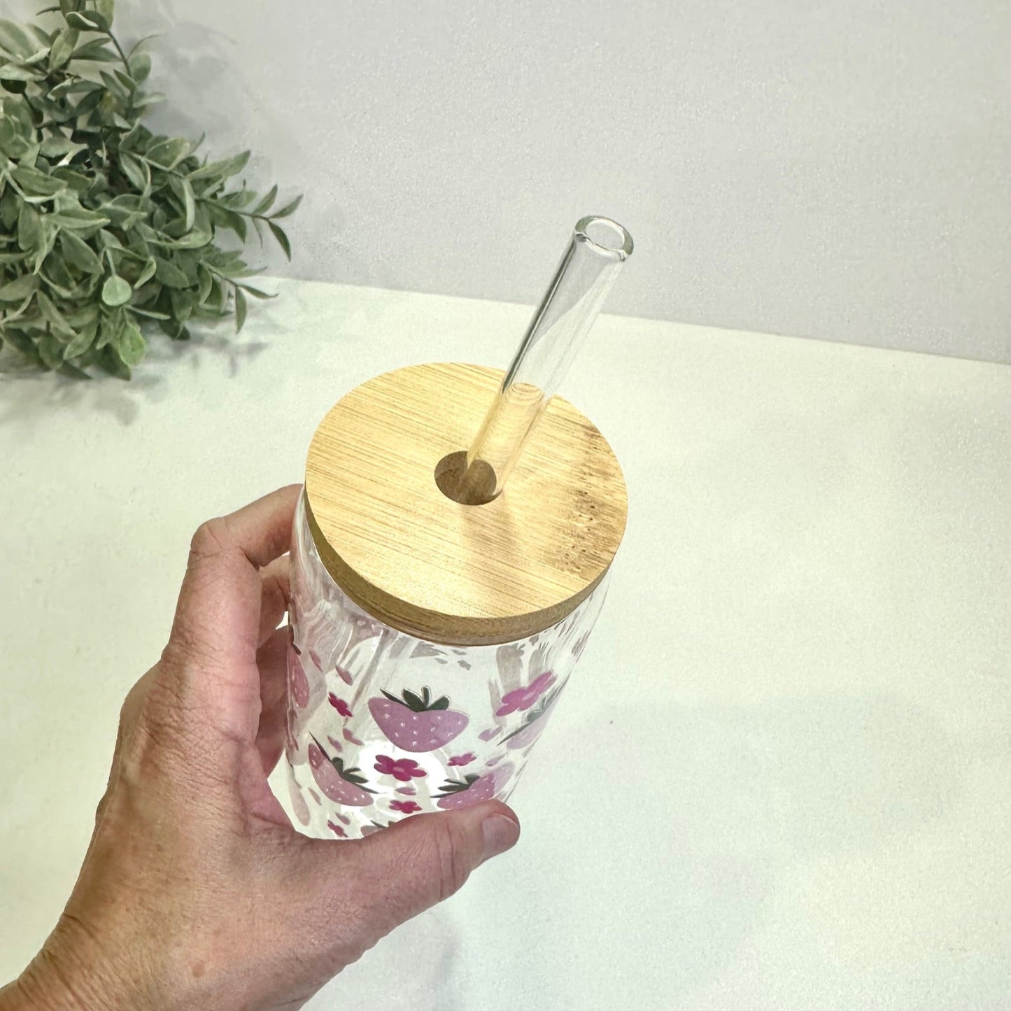 Hand holding a clear tumbler with floral design and wooden lid on a light surface.