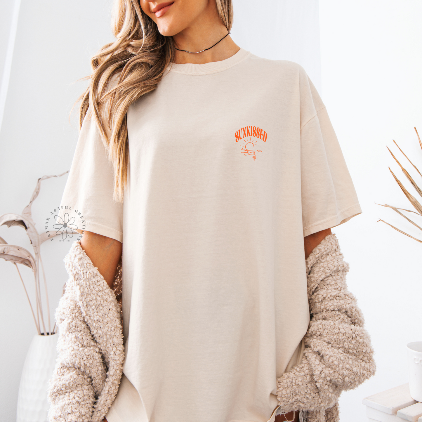 A person wearing a ivory oversized unisex t-shirt with the text 'SUNKISSED' and a graphic pocket design in orange.