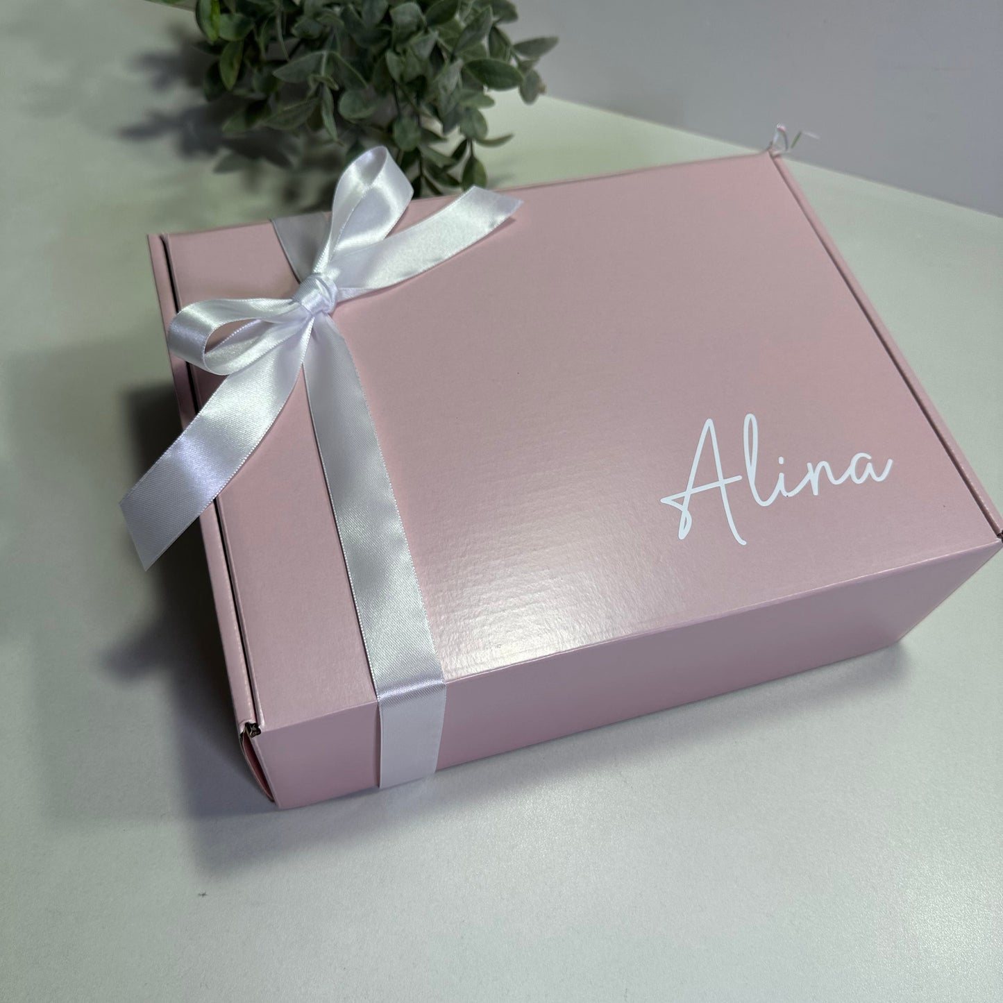 Pink personalized gift box with a white ribbon on a light surface with a plant in the corner