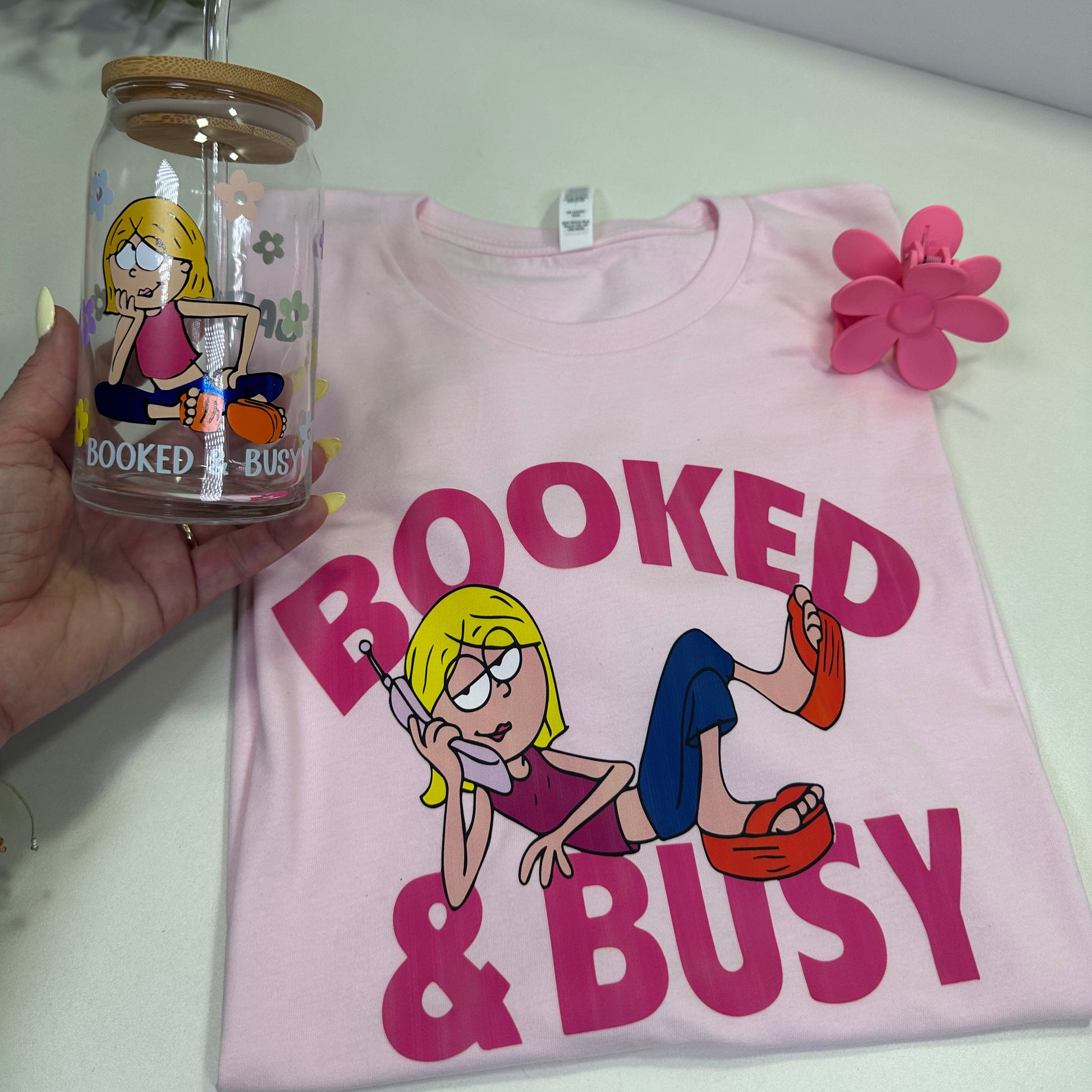 pink booked and busy shirt, pink flower hair clip and hand holding a glass can cup with retro flowers and lizzie McGuire seat and under booked and busy words