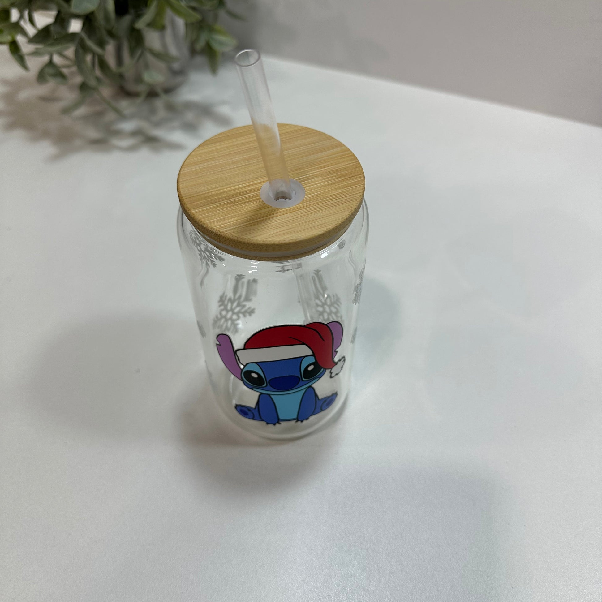 Glass tumbler with wooden lid and straw, featuring a Christmas stitch character, on a white surface.