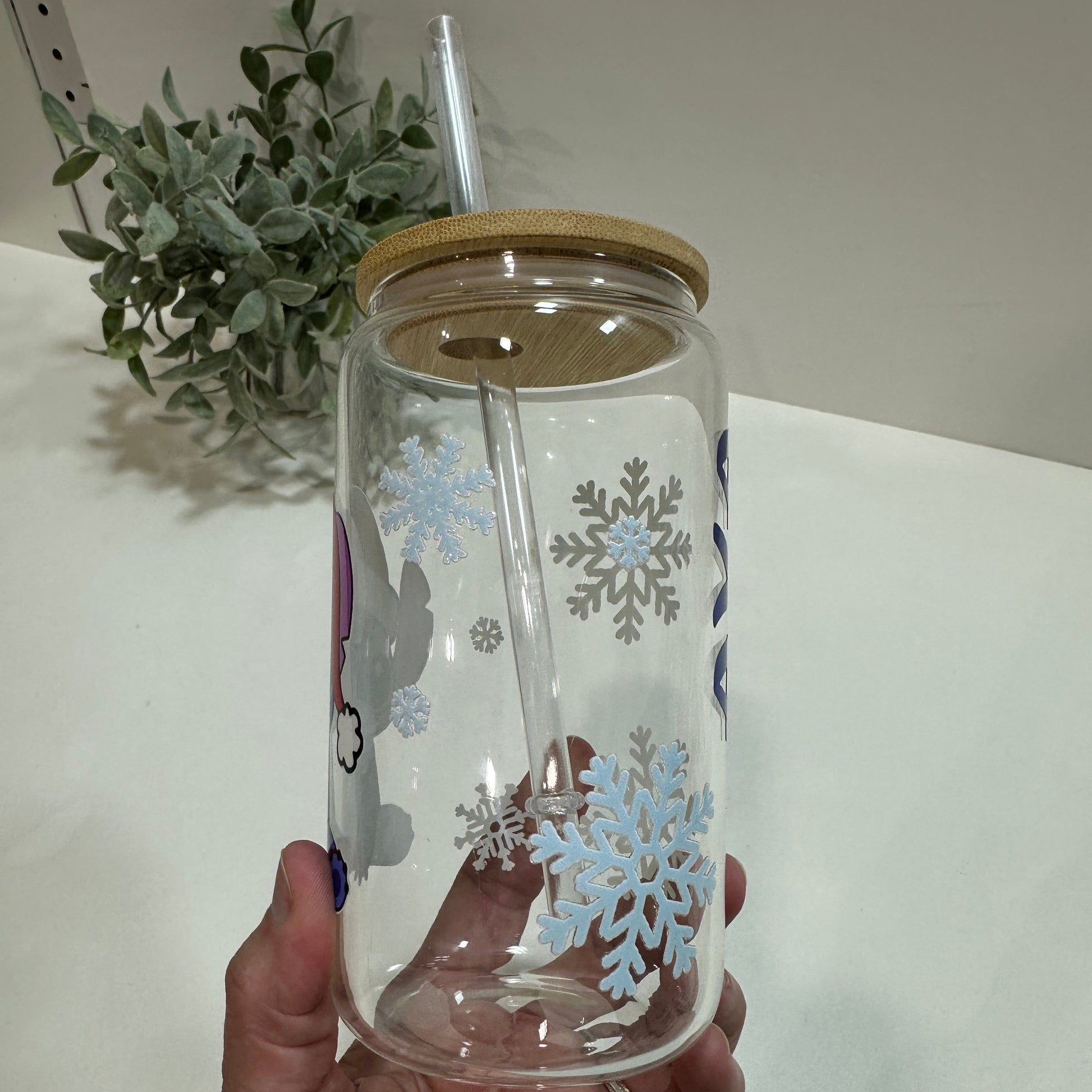 Clear tumbler with snowflake design held by a hand, with a plant in the background.