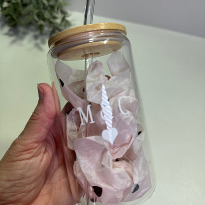 Clear glass tumbler with M & C and a heart design and straw held by a hand against a light background