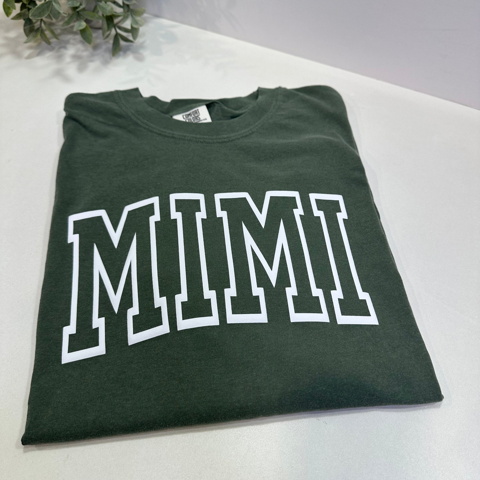 Green t-shirt with 'MIMI' printed on it, placed on a white surface.