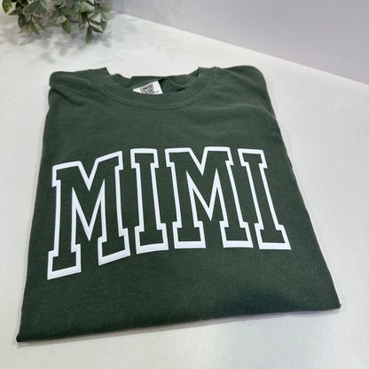 Green t-shirt with 'MIMI' printed on it, placed on a white surface.