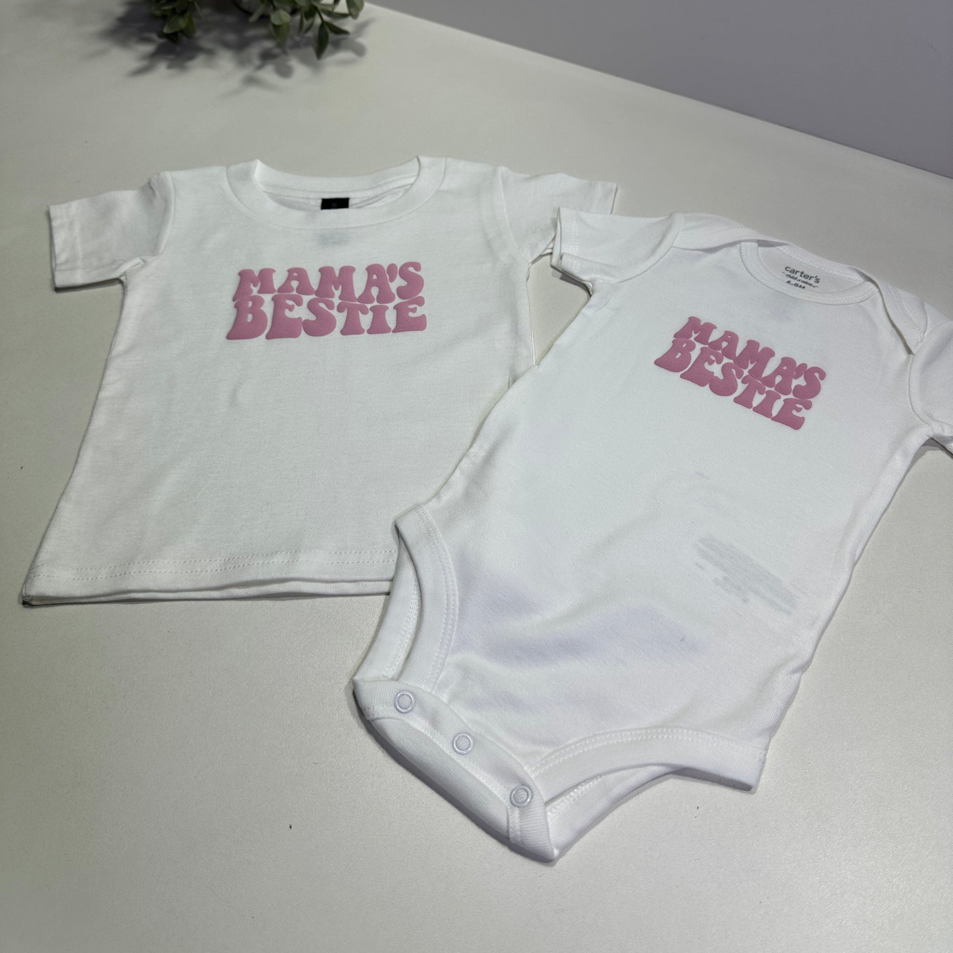 White baby onesie and kid shirt with pink "mama's bestie" text on a light gray surface.