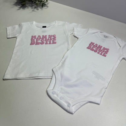 White baby onesie and kid shirt with pink "mama's bestie" text on a light gray surface.