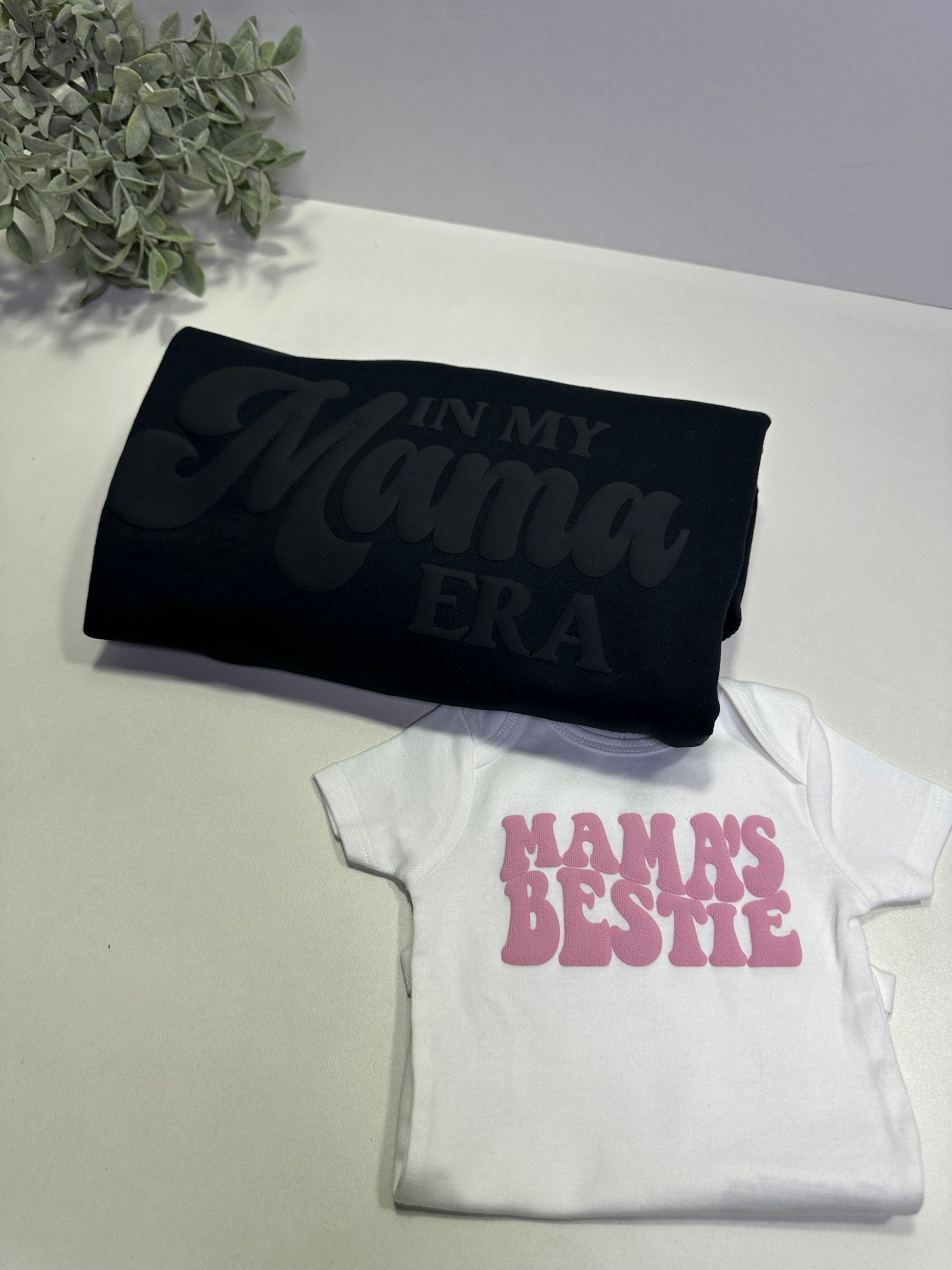 One folded black sweatshirt with "In my Mama Era" text in black and white baby onesie with pink text "mama's bestie"  on a light surface
