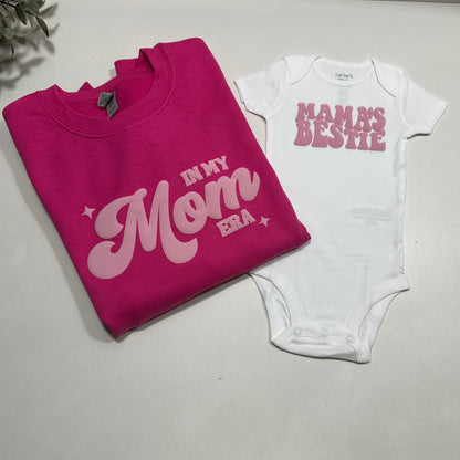 White onesie with pink text "mama's bestie" and dark pink sweatshirt with light pink text "in my mom era" on a white background