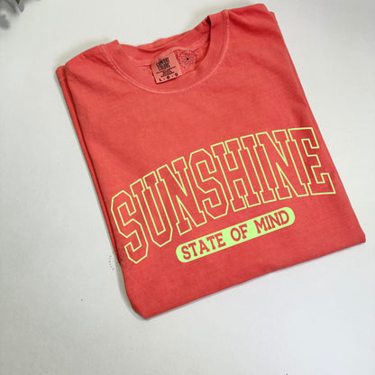 Folded neon orange t-shirt with bright yellow text "sunshine state of mind" on a white surface