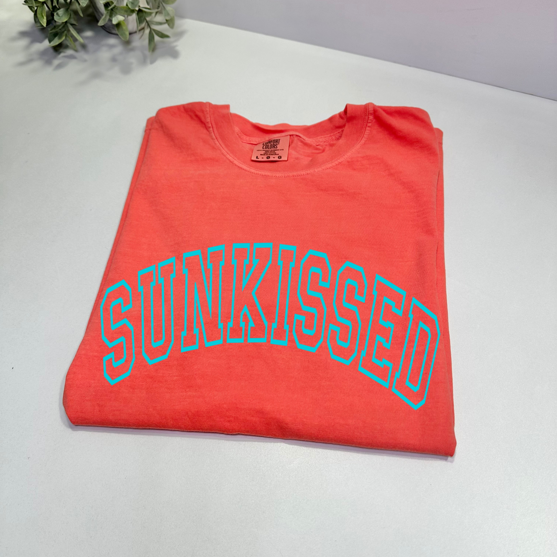 A  bright salmon Comfort Colors T-shirt with the word 'SUNKISSED' printed in bold sky blue puff print.