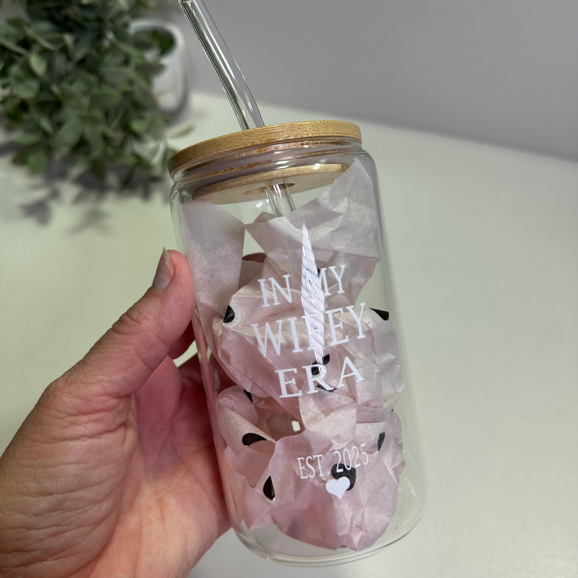 Hand holding a clear glass tumbler  with in my wifey era custom design and straw, against a neutral background.