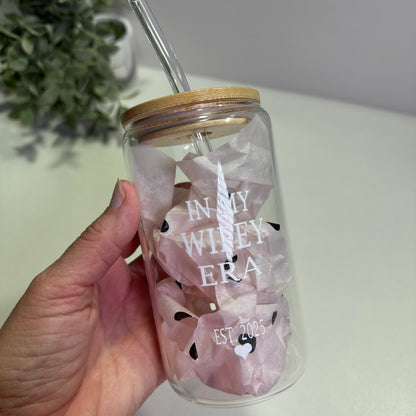 Hand holding a clear glass tumbler  with in my wifey era custom design and straw, against a neutral background.
