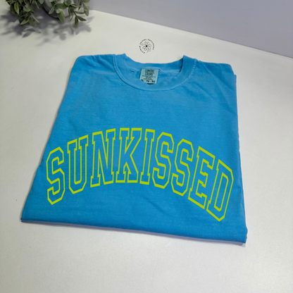 A sapphire Comfort Colors T-shirt with the word 'SUNKISSED' printed in bold neon yellow puff print.