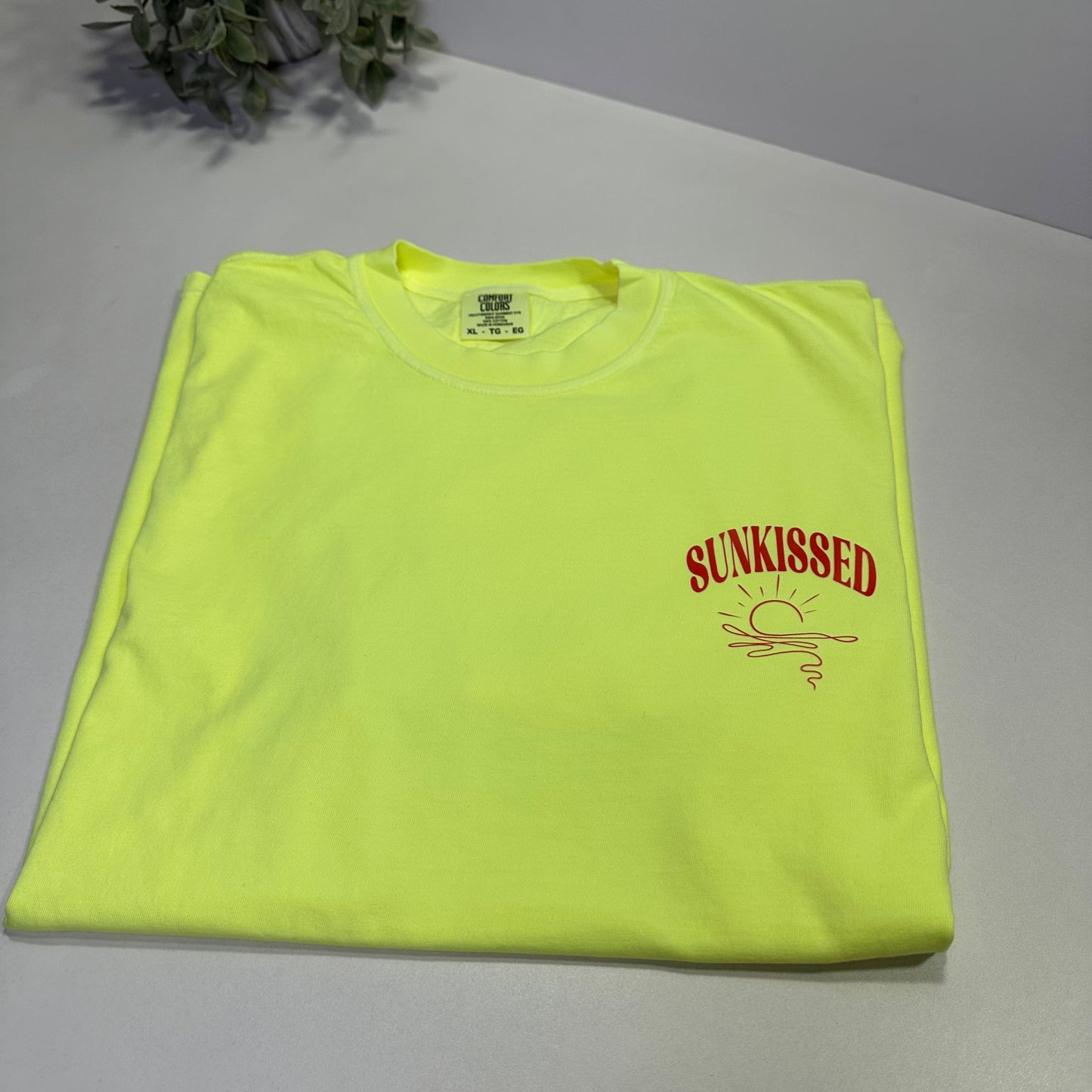 neon lemon t-shirt with orange-red text on a white surface next to a plant