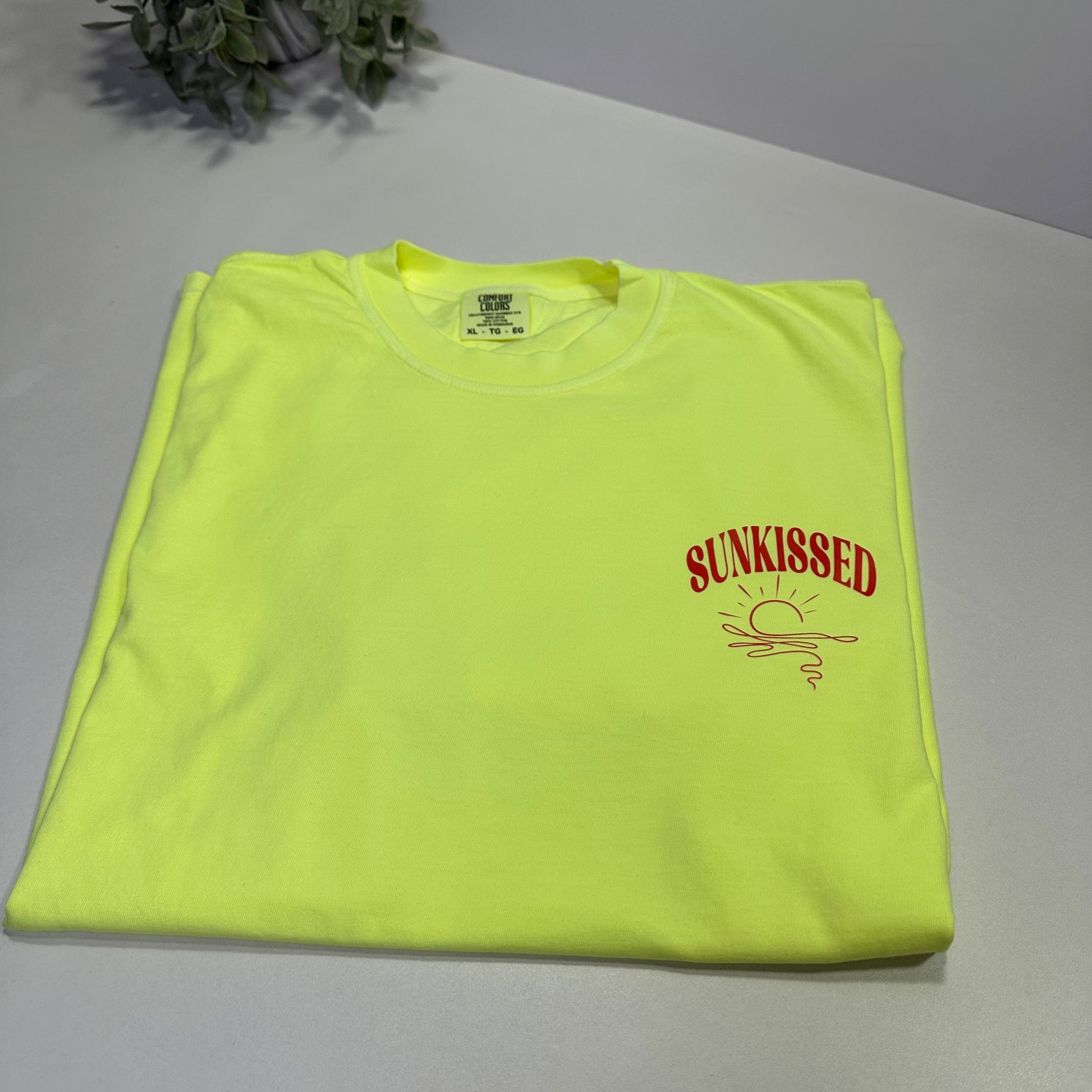 neon lemon t-shirt with orange-red text on a white surface next to a plant