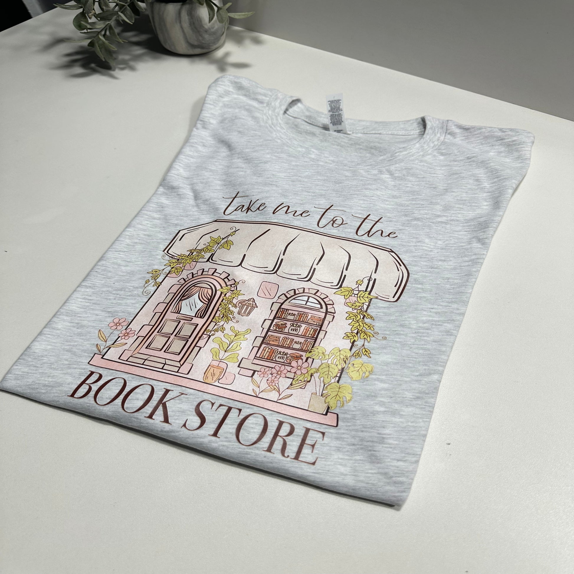 Light gray t shirt with an aesthetic library graphic design that said take me to the bookstore