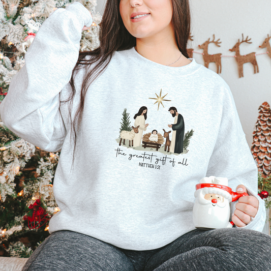 A woman sitting in front of a Christmas tree wearing a gray sweatshirt with a nativity scene design.